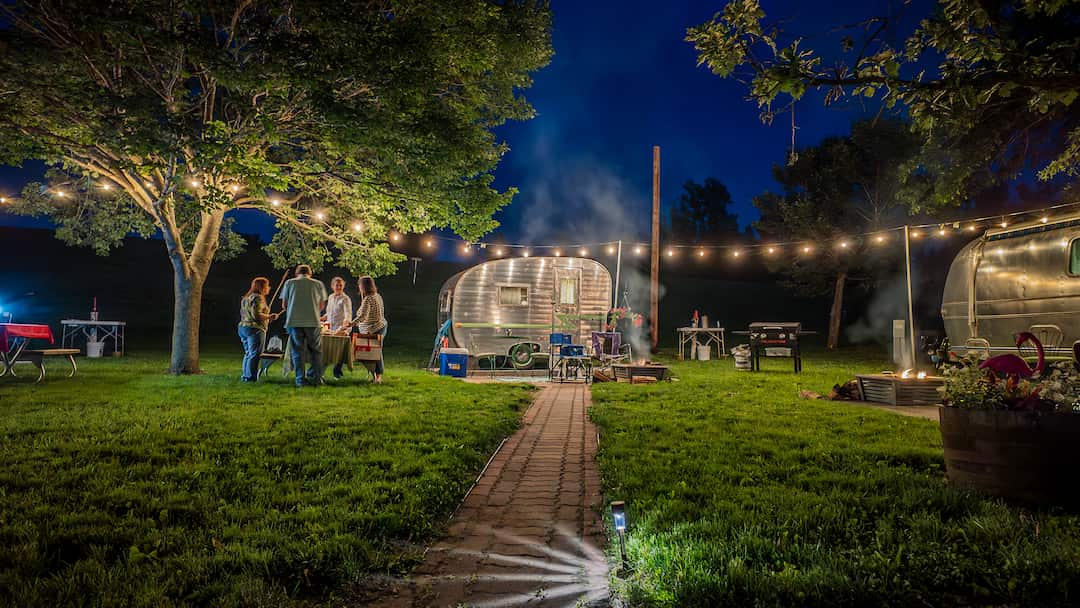 Friends walking toward a vintage camper under string lights at dusk at Alta Campground in Newton, Iowa